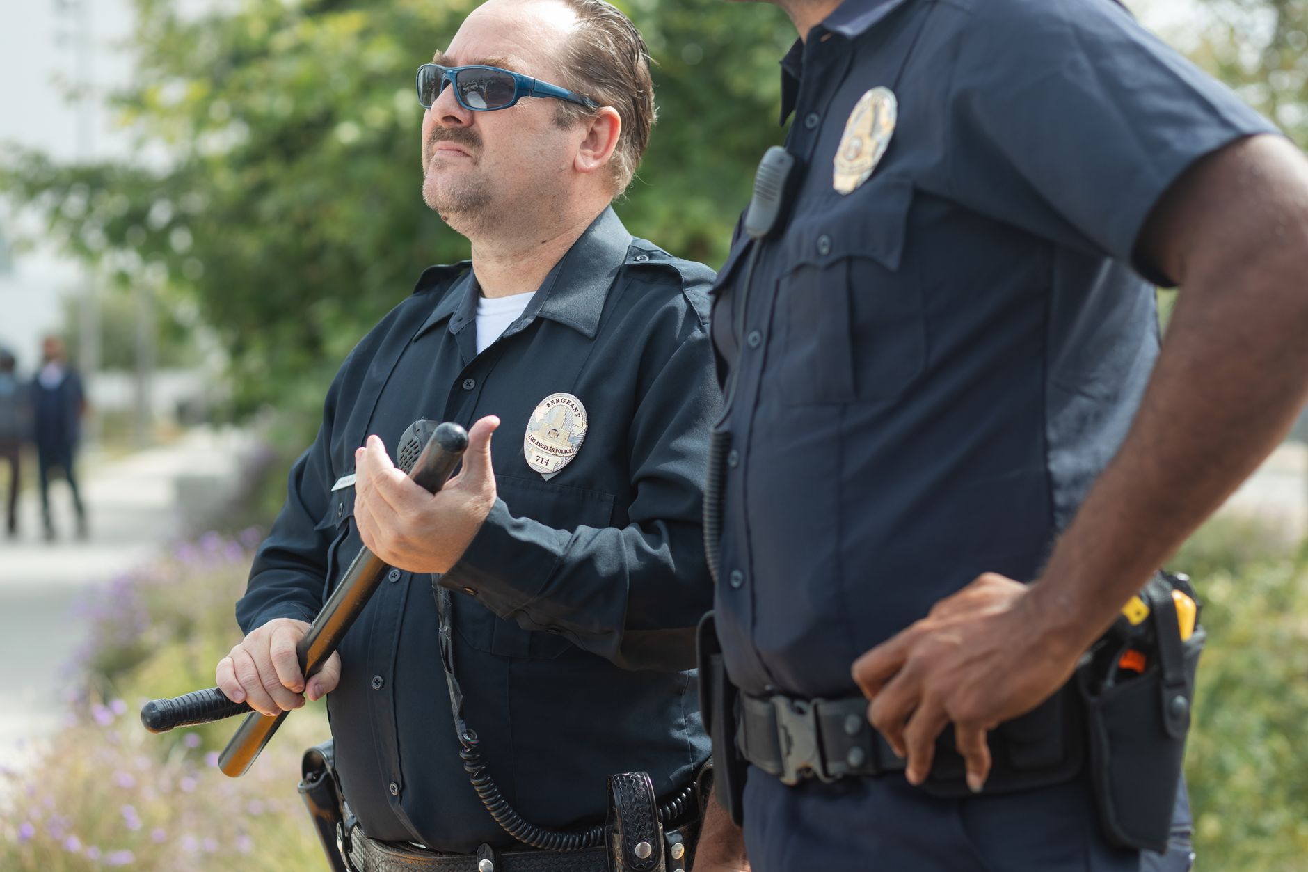 Two uniformed police officers stand outdoors. One officer, wearing sunglasses, holds a wooden baton across his body, while the other stands beside him with a hand resting near his duty belt.