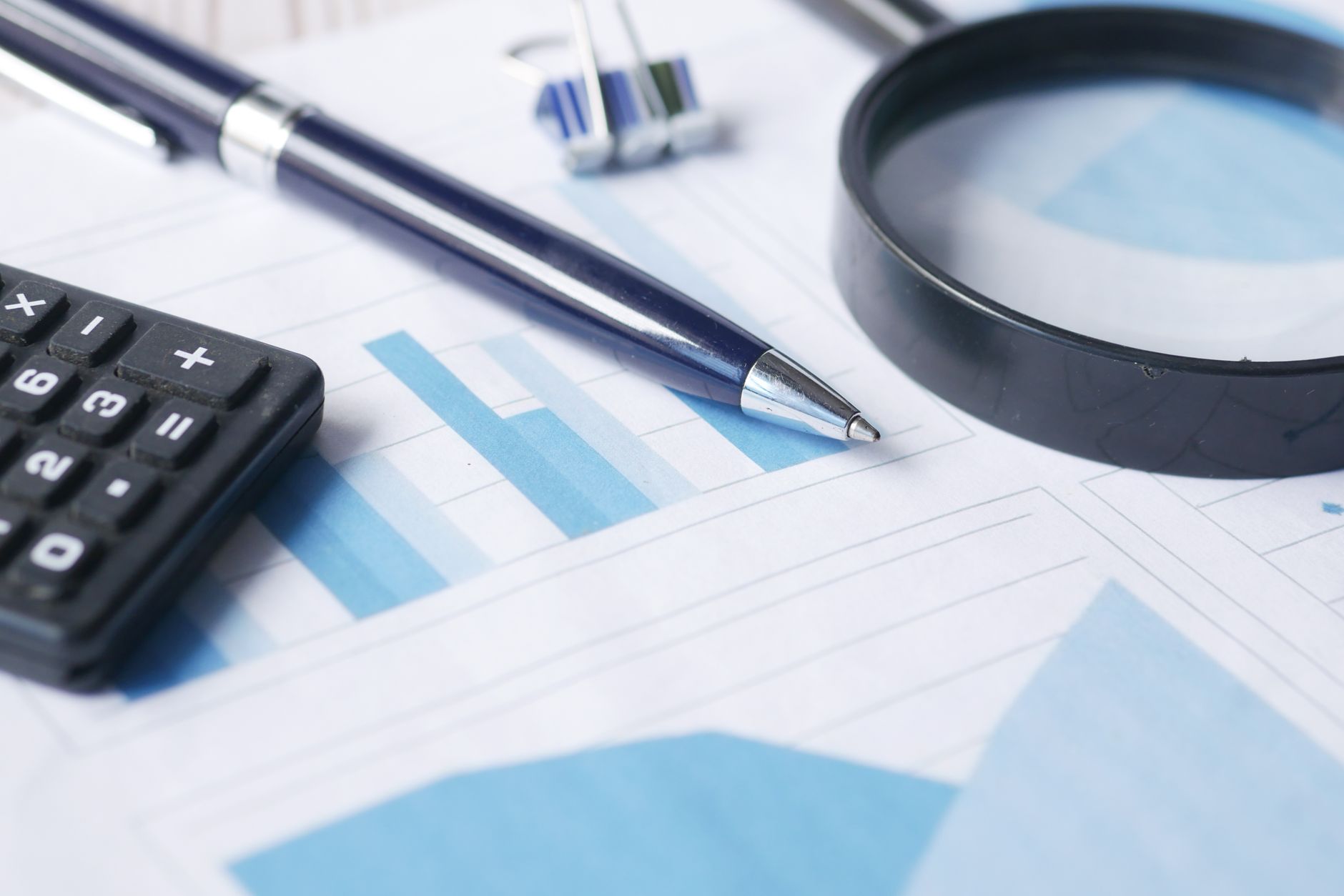 Close-up of a calculator, pen, magnifying glass, and eraser resting on financial documents featuring blue bar and line charts.